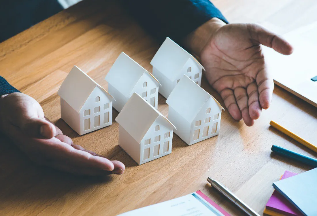 Hands framing a row of miniature house models on a desk, symbolising property portfolio growth and the shift from hands-on management to strategic investment.