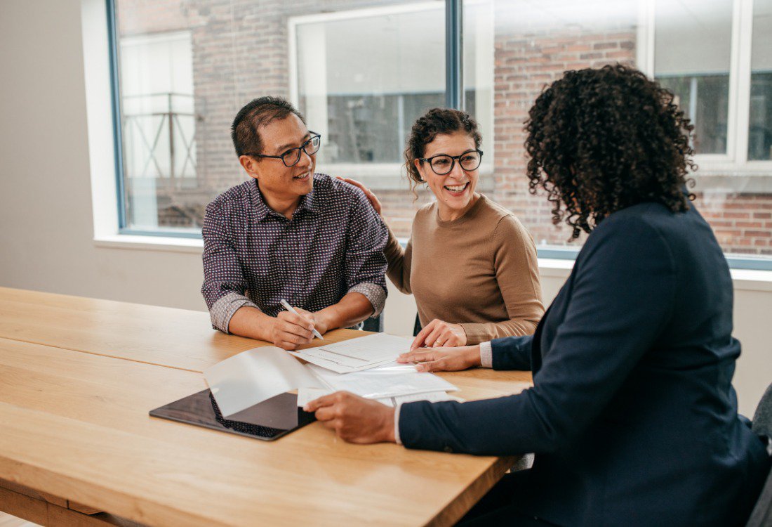A property manager discusses options for managing a property portfolio with a smiling couple, pointing to documents on a table.