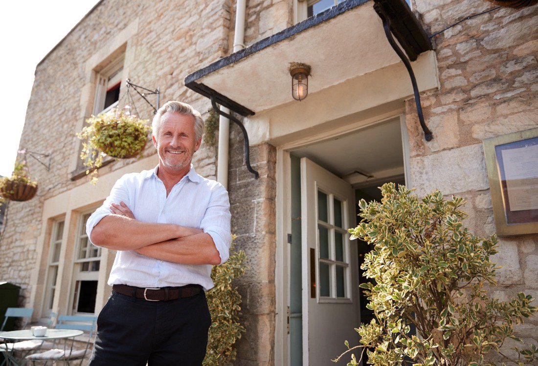 A confident landlord with his arms crossed stands smiling in front of a house, representing successful property portfolio optimisation.